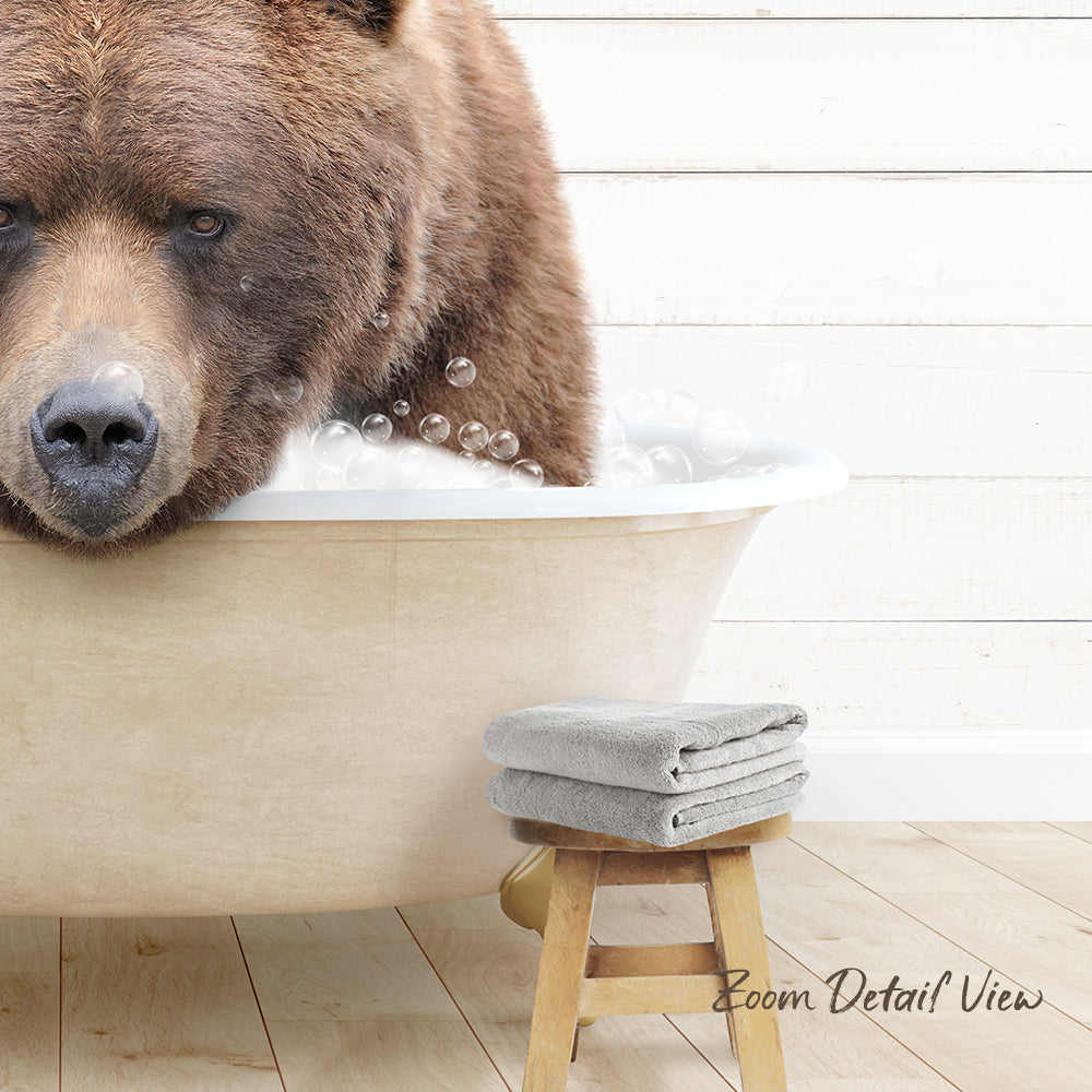 A large brown bear is sitting in a bathtub filled with bubbles, with a towel and stool nearby.