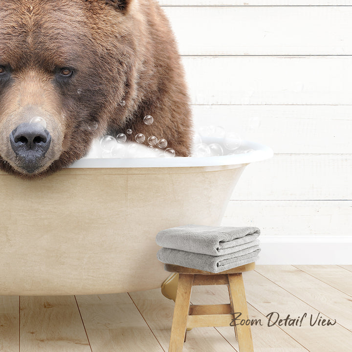 A large brown bear is sitting in a bathtub filled with bubbles, with a towel and stool nearby.