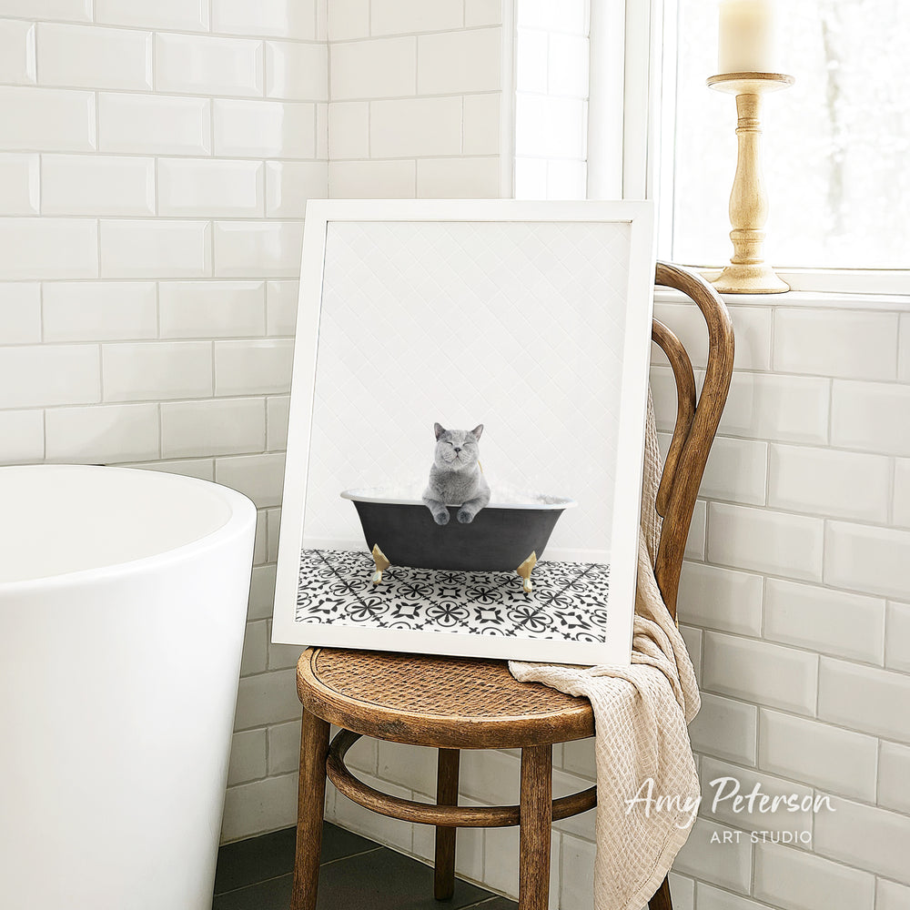 A framed photograph of a gray cat sitting in a bathtub, with a wooden chair and a white tiled wall in the background.