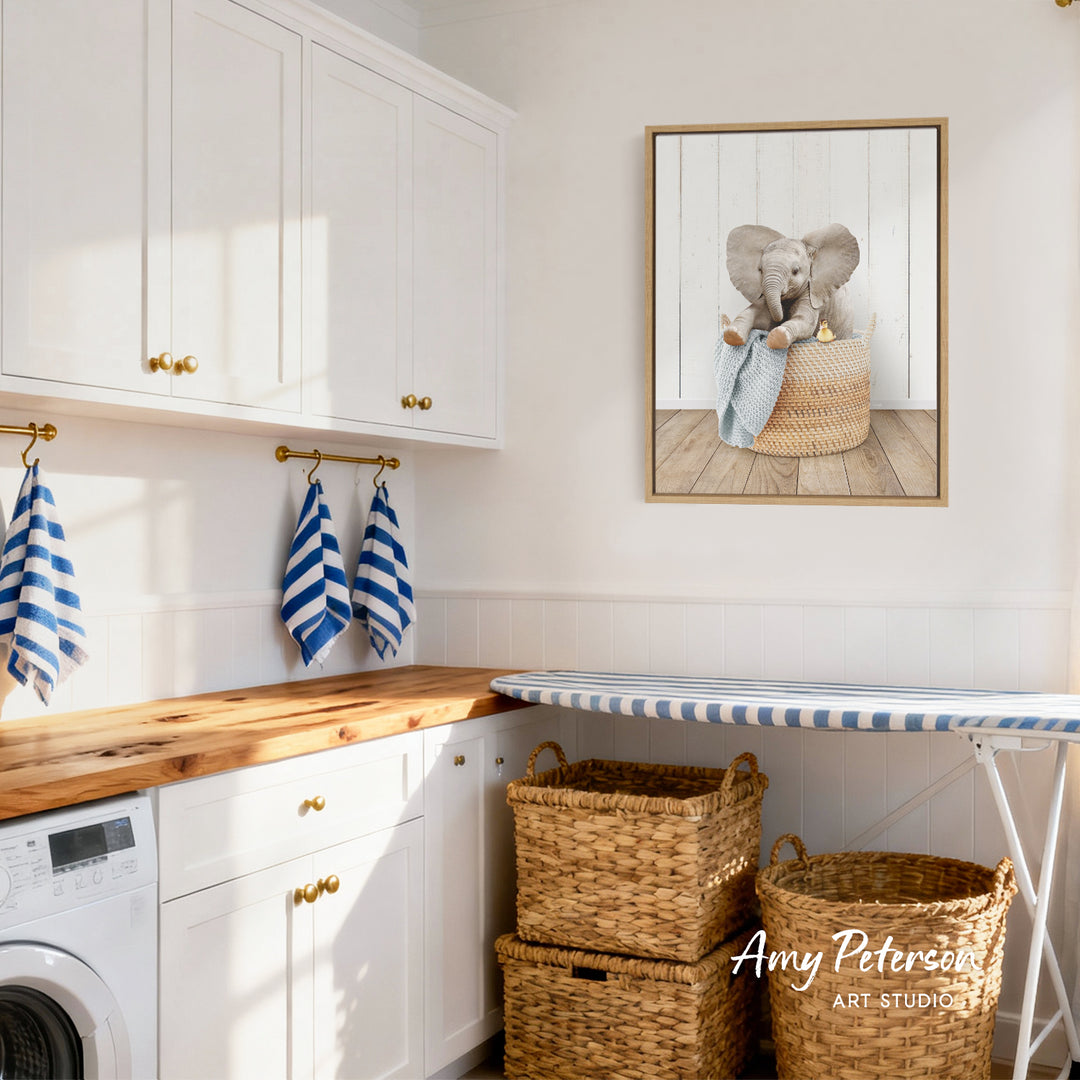 A cozy laundry room with white cabinets, a wooden countertop, and a framed picture of an elephant sitting in a basket. There are also two woven baskets and a striped ironing board in the room.