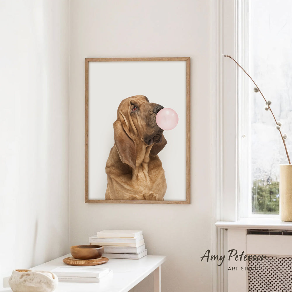 A framed photograph of a dog blowing a bubble gum bubble is hanging on a white wall in a room with a white table and a window.