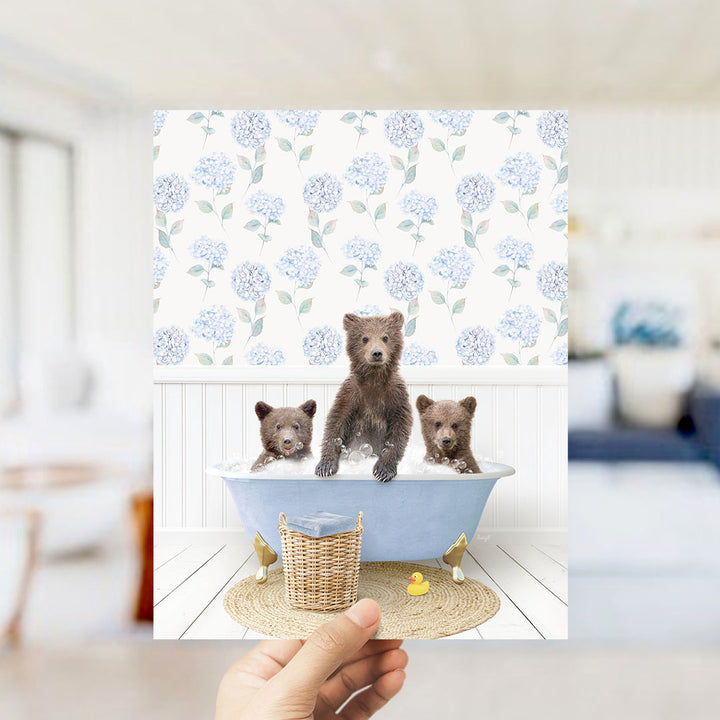 A hand holds up a framed image depicting three adorable brown bears sitting in a blue bathtub, surrounded by a floral patterned wall.
