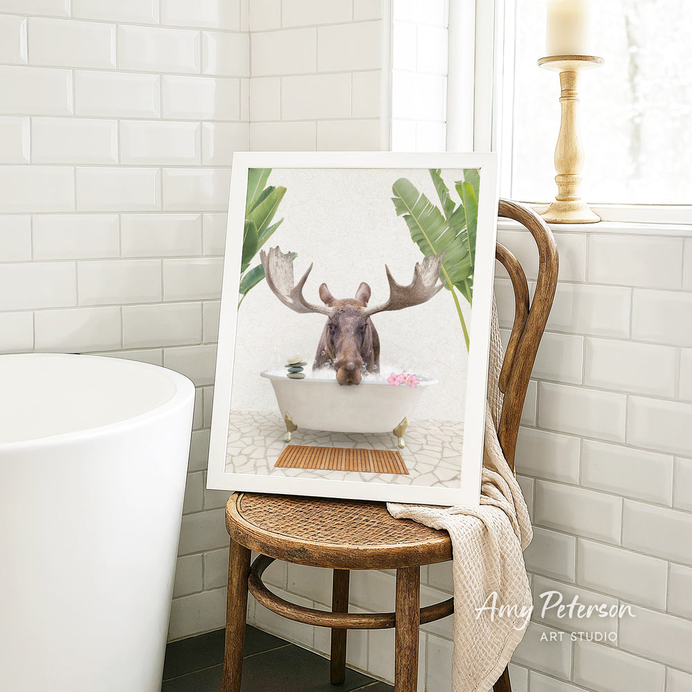 A framed image of a moose sitting in a bathtub, surrounded by tropical plants, is displayed on a wooden chair in a bathroom setting.
