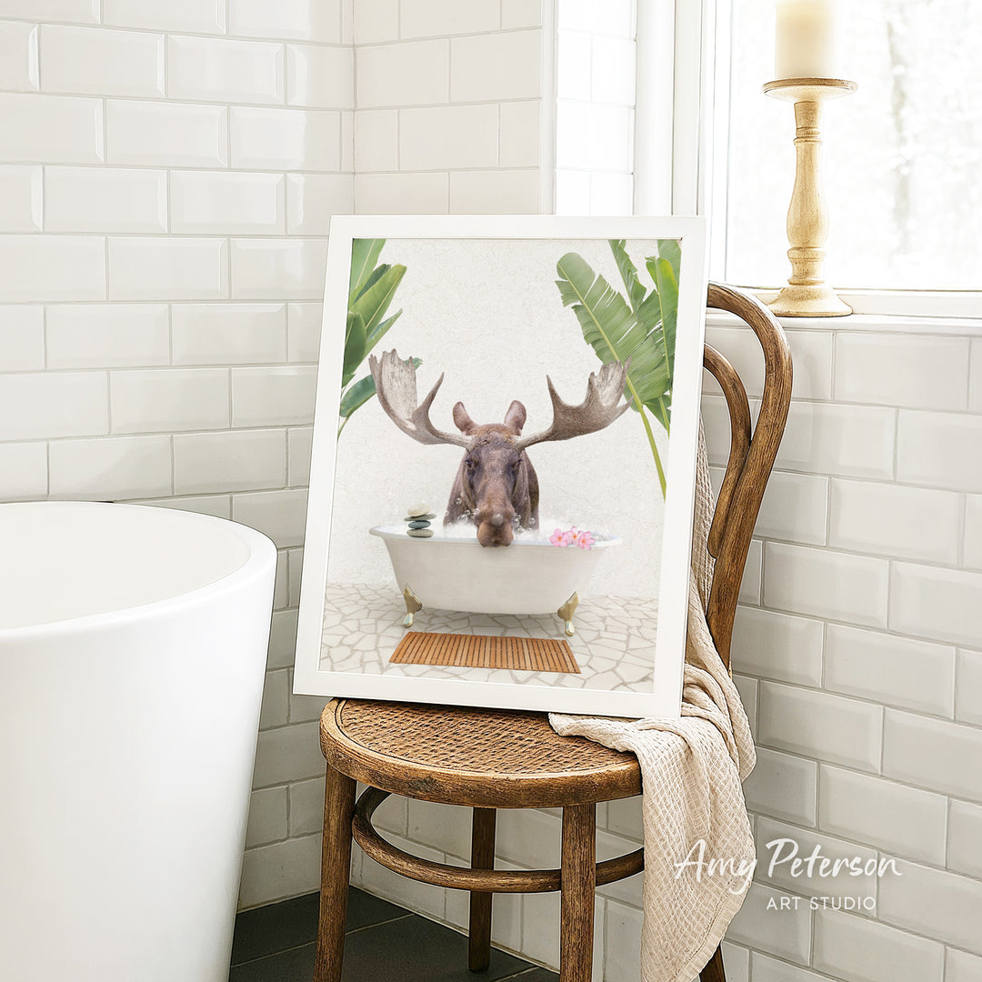 A framed image of a moose sitting in a bathtub, surrounded by tropical plants, is displayed on a wooden chair in a bathroom setting.