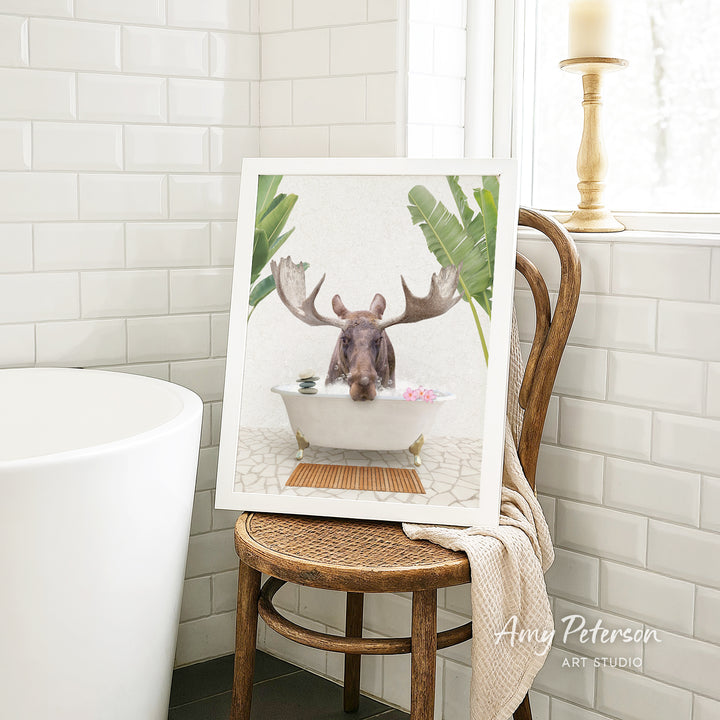A framed image of a moose sitting in a bathtub, surrounded by tropical plants, is displayed on a wooden chair in a bathroom setting.