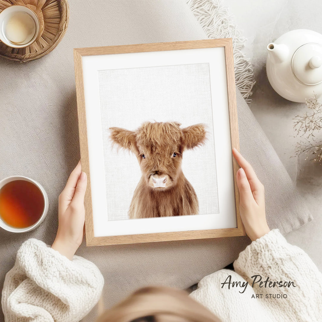 A person is holding a framed picture of a brown cow, which is the main focus of the image. The person is sitting on a couch, and there are other objects in the scene, such as a teapot, a cup, and a bowl.