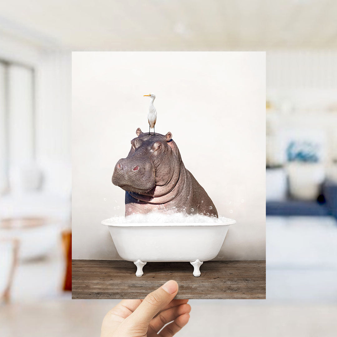 A hand holds up a framed image of a hippopotamus sitting in a bathtub, with a seagull perched on its head.