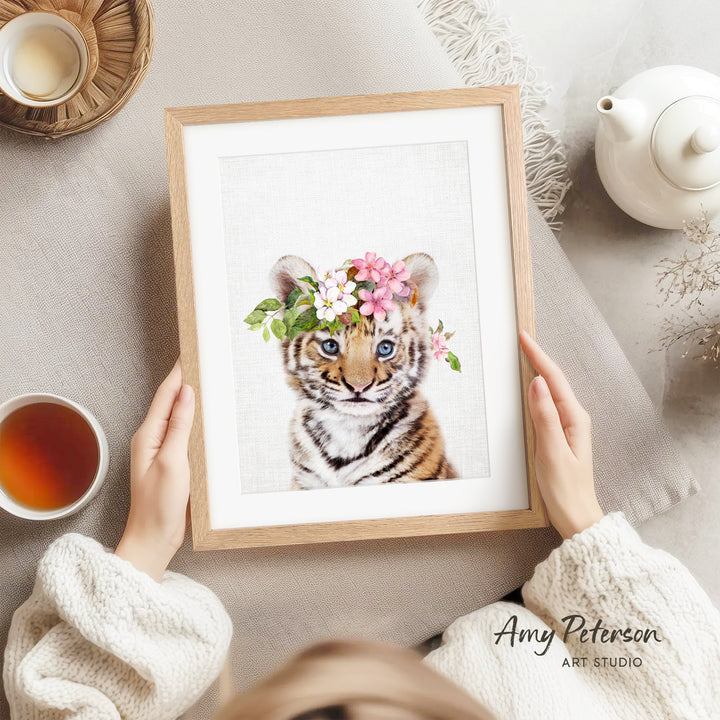 A person is holding a framed picture of a tiger wearing a flower crown, with a teapot and a cup of tea nearby.