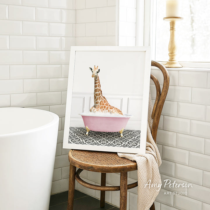 A framed image of a giraffe sitting in a pink bathtub is displayed on a wooden chair in a bathroom setting.