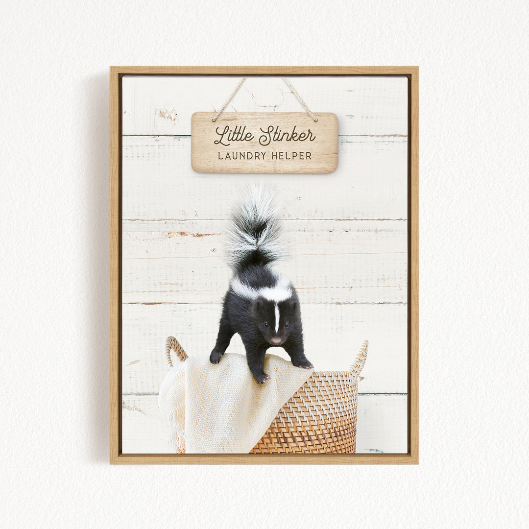 A black and white skunk standing on a woven basket, with a wooden sign above that reads "Little Stinker Laundry Helper".