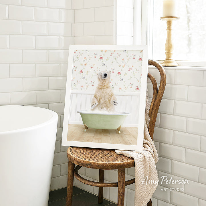 A framed image of a polar bear sitting in a bathtub, surrounded by a floral pattern, is displayed on a wooden chair in a bathroom setting.