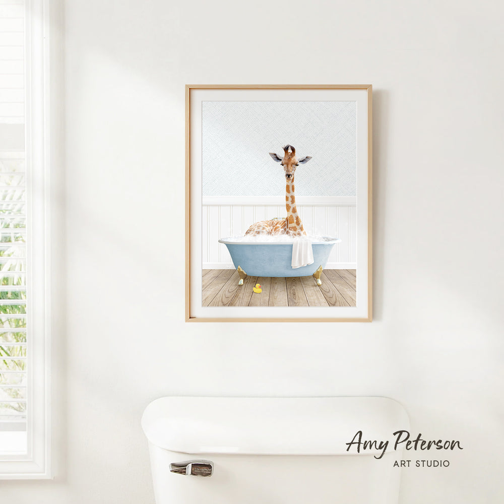 A framed print of a giraffe sitting in a bathtub is hanging on a white wall.