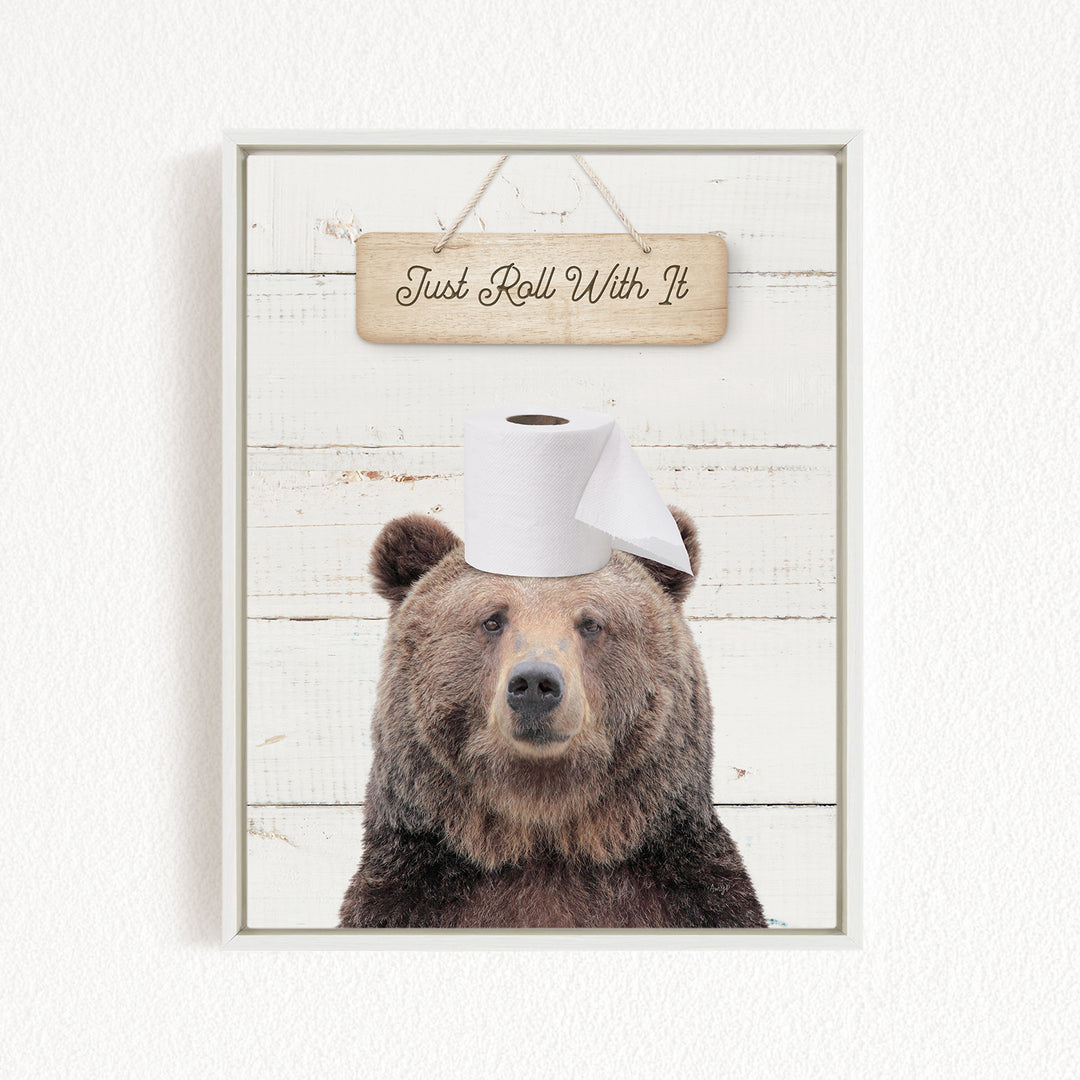 A wooden sign with the text "Just Roll With It" hangs above a picture of a bear wearing a toilet paper roll on its head.