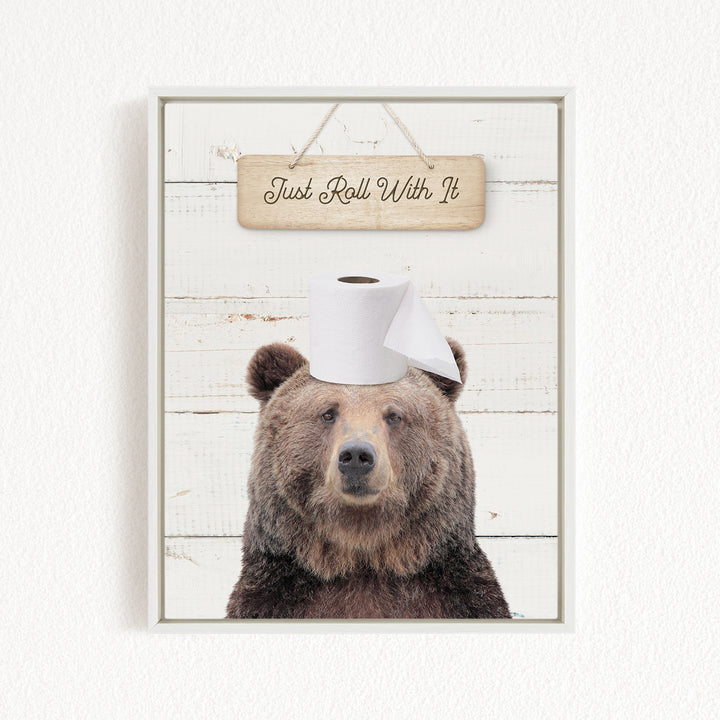 A wooden sign with the text "Just Roll With It" hangs above a picture of a bear wearing a toilet paper roll on its head.