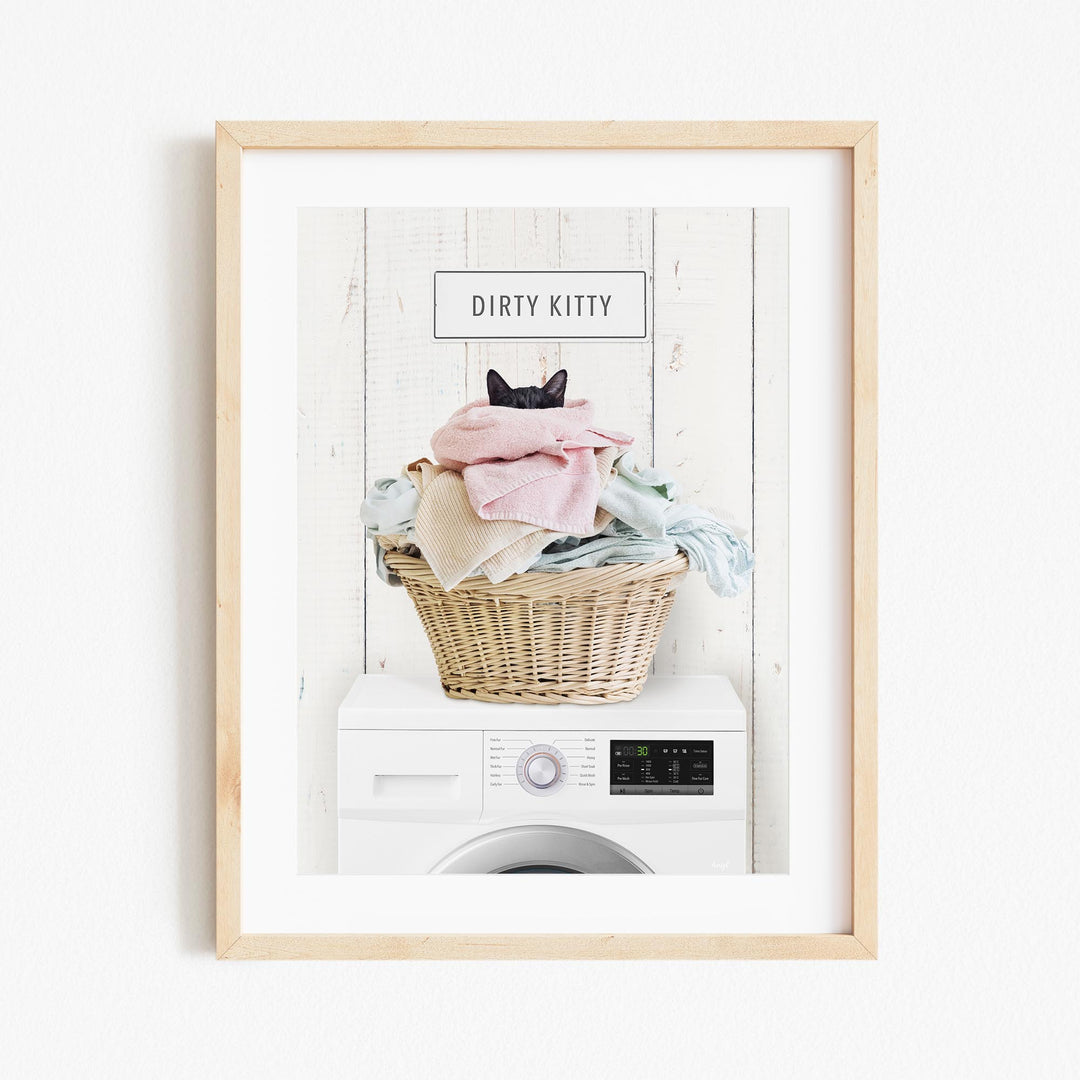 A framed image depicting a cat sitting on top of a basket filled with laundry, with the text "Dirty Kitty" displayed above the basket.