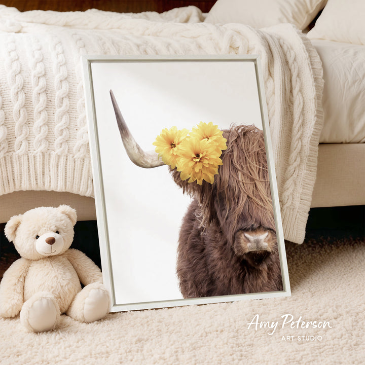 A framed photograph of a brown cow with horns and yellow flowers on its head, placed on a white blanket on a bed next to a teddy bear.