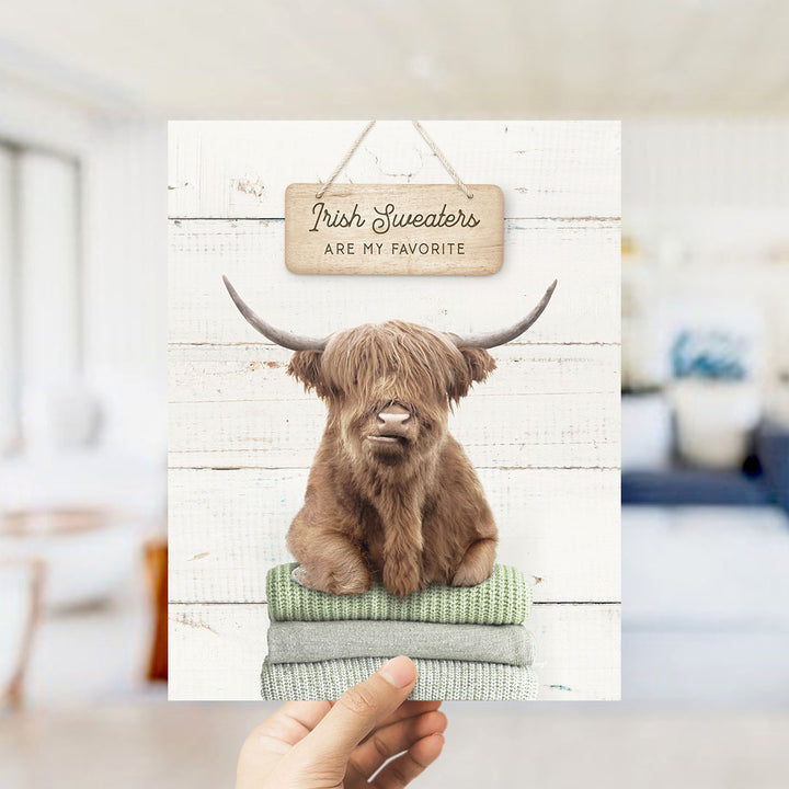 A hand is holding up a framed print featuring a brown Highland cow with horns, sitting on a stack of green sweaters. The text above the cow reads "Irish Sweaters are my favorite".