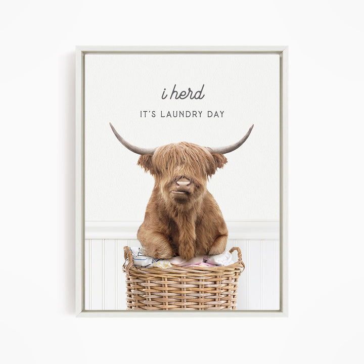 A brown Highland cow with large horns sits in a wicker basket, with the text "It's Laundry Day" above the image.