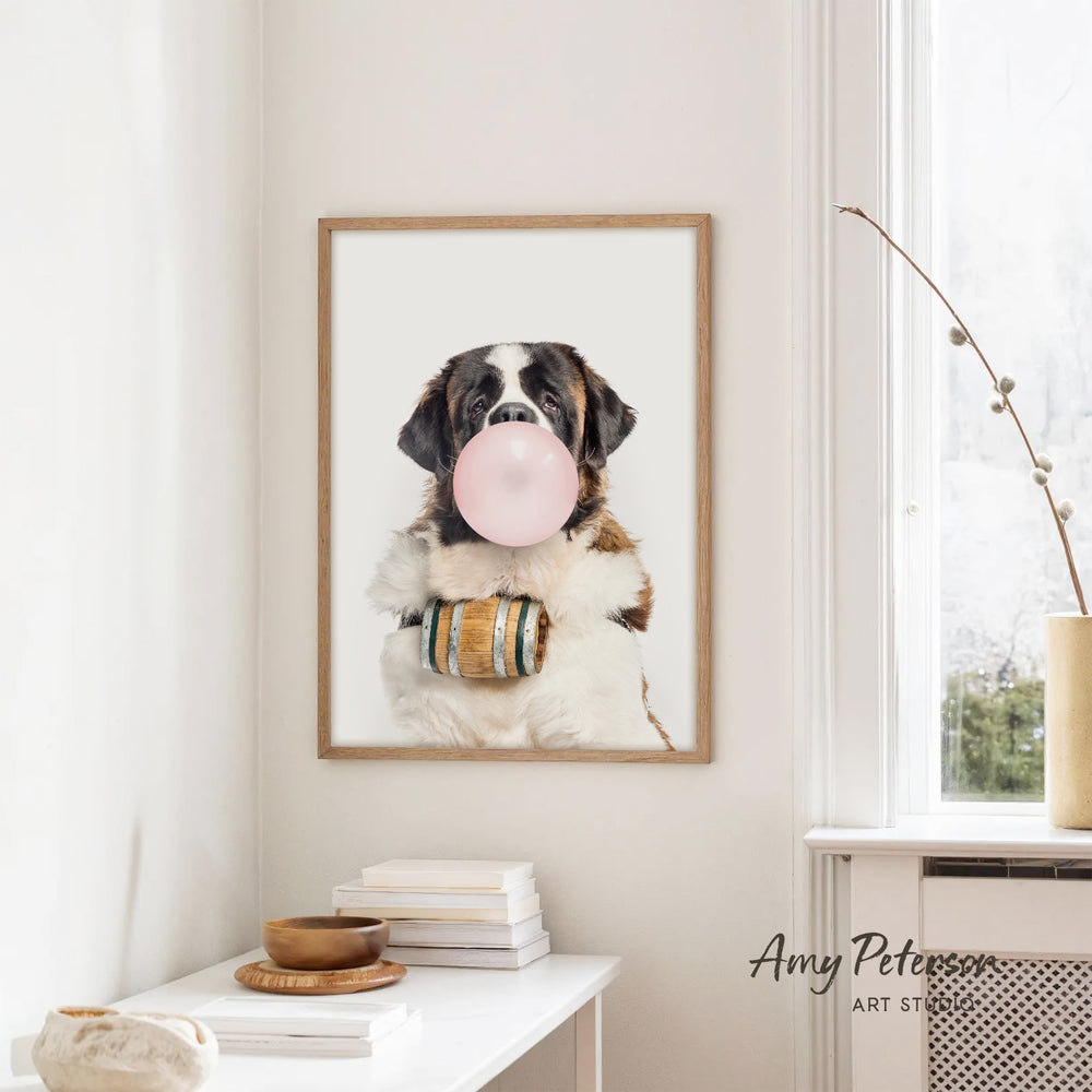 A framed photograph of a Saint Bernard dog blowing a bubble with a pink bubble gum bubble, sitting on a white table with a stack of books and a small plant.