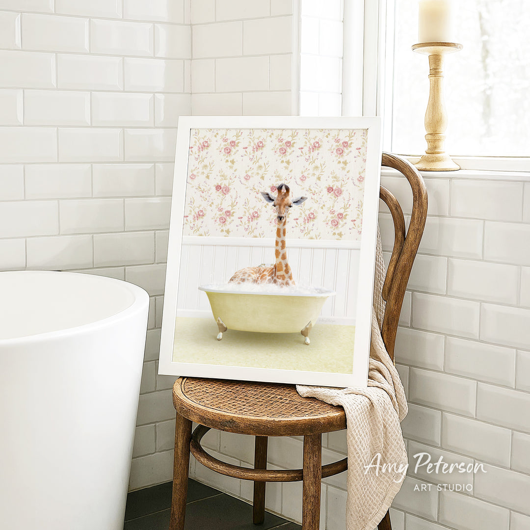 A framed image of a giraffe sitting in a bathtub, surrounded by a floral pattern, is displayed on a wooden chair in a bathroom setting.