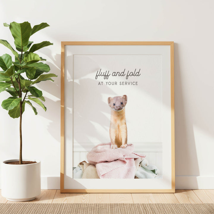 A framed photograph of a small animal, likely a cat, sitting on a pile of clothes in a room with a potted plant to the left.