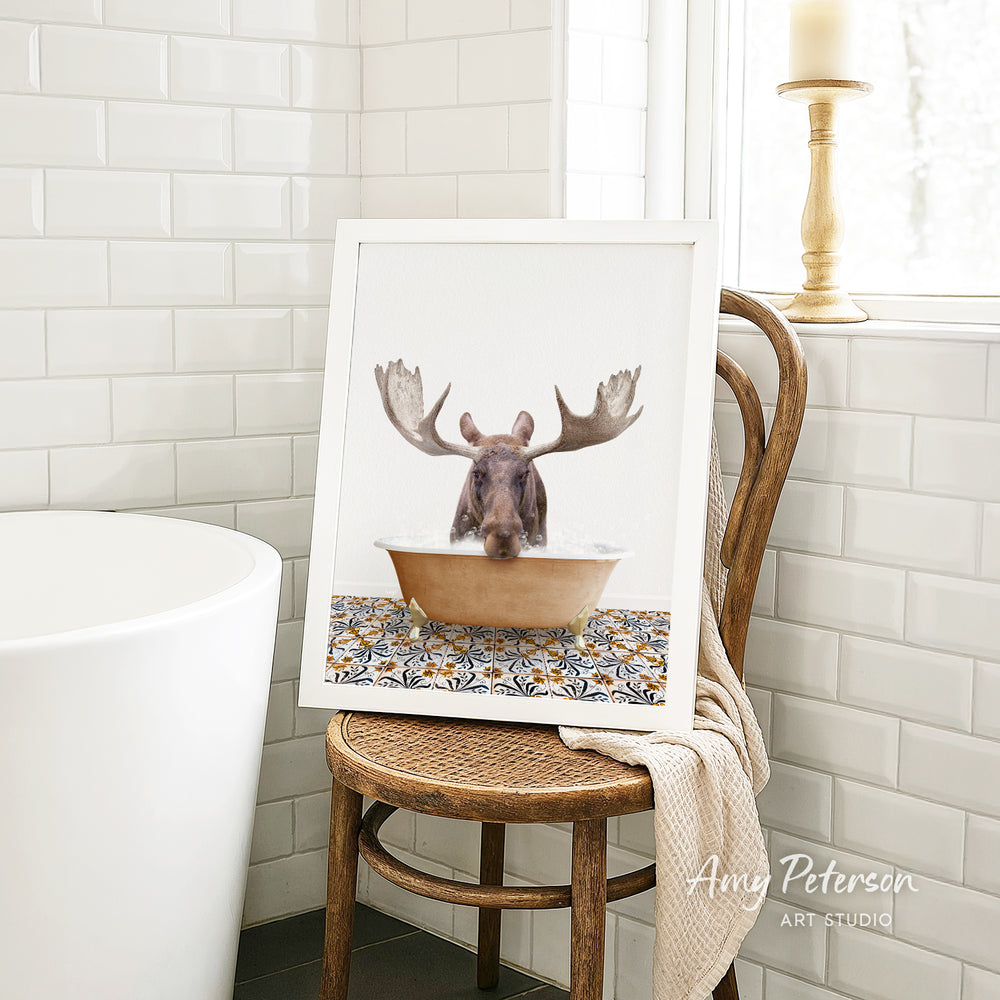 A framed image of a moose sitting in a bathtub, with its antlers raised, is displayed on a wooden chair in a bathroom setting.