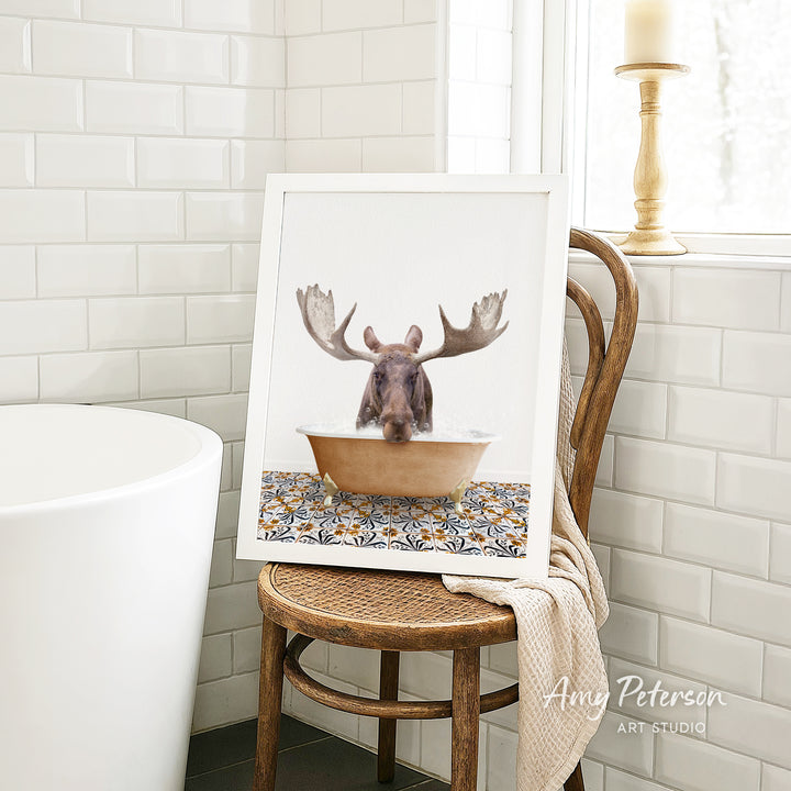 A framed image of a moose sitting in a bathtub, with its antlers raised, is displayed on a wooden chair in a bathroom setting.