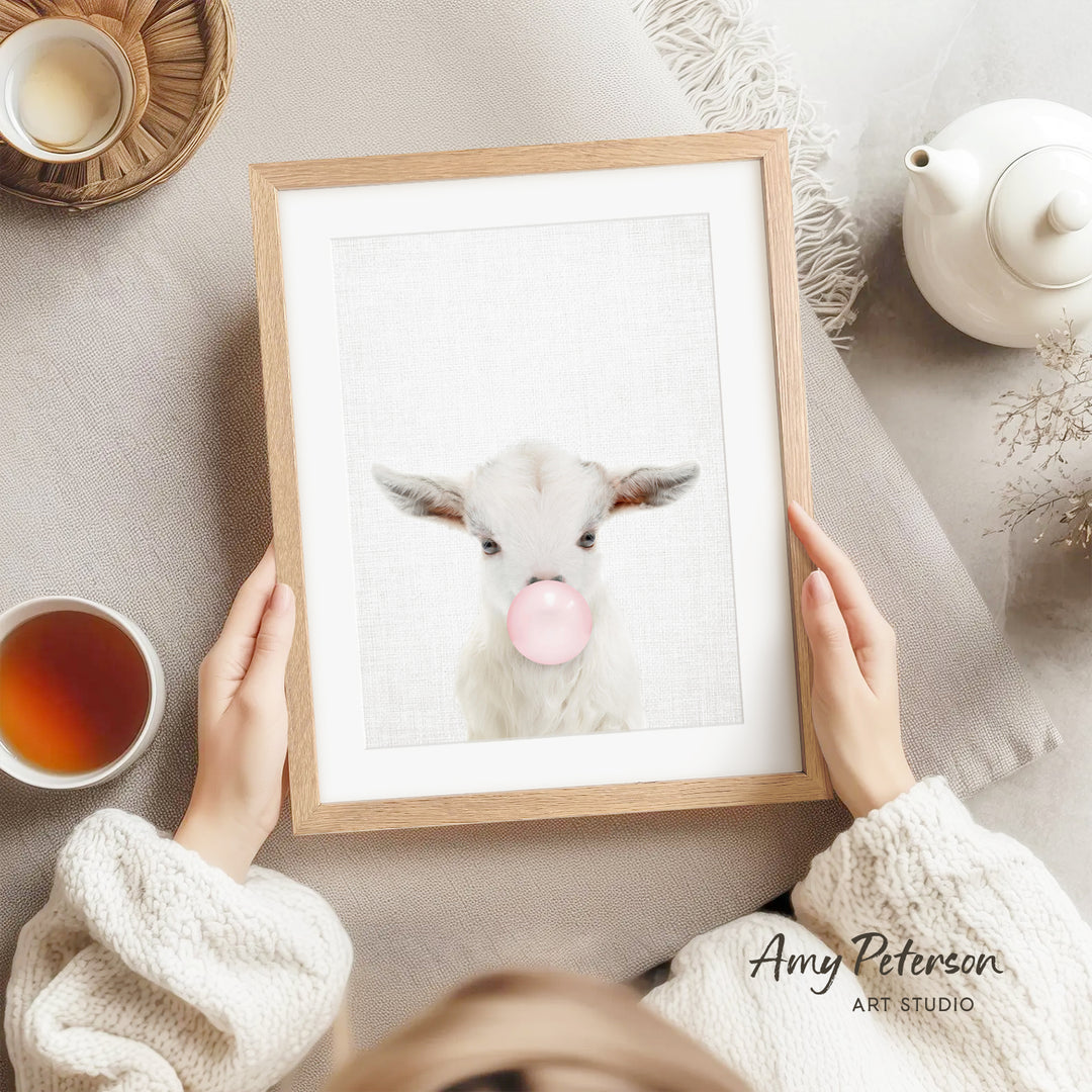 A person is holding a framed picture of a white goat blowing a bubble with a pink bubblegum bubble.