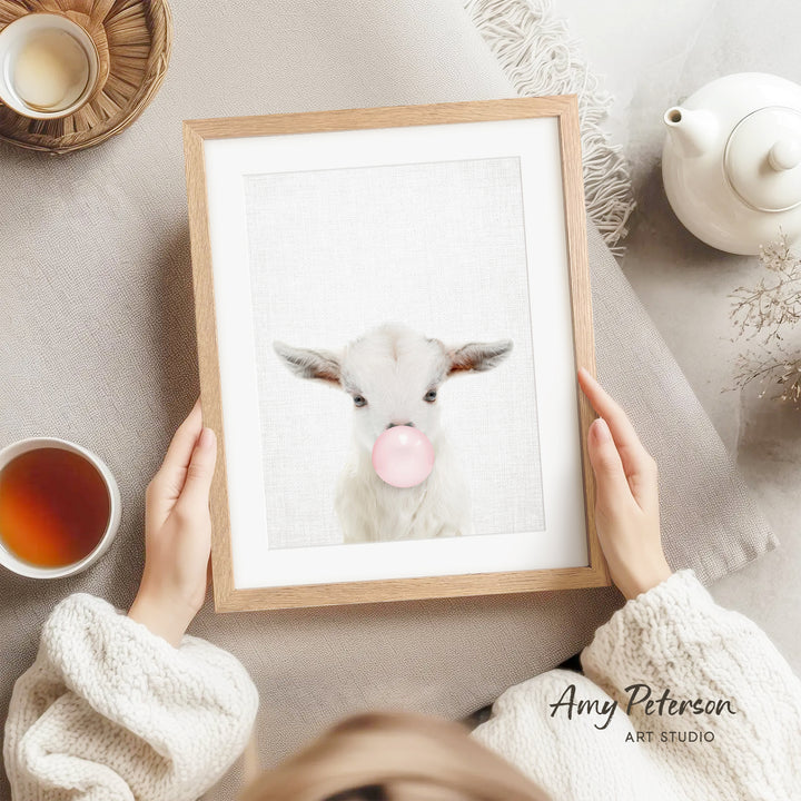 A person is holding a framed picture of a white goat blowing a bubble with a pink bubblegum bubble.