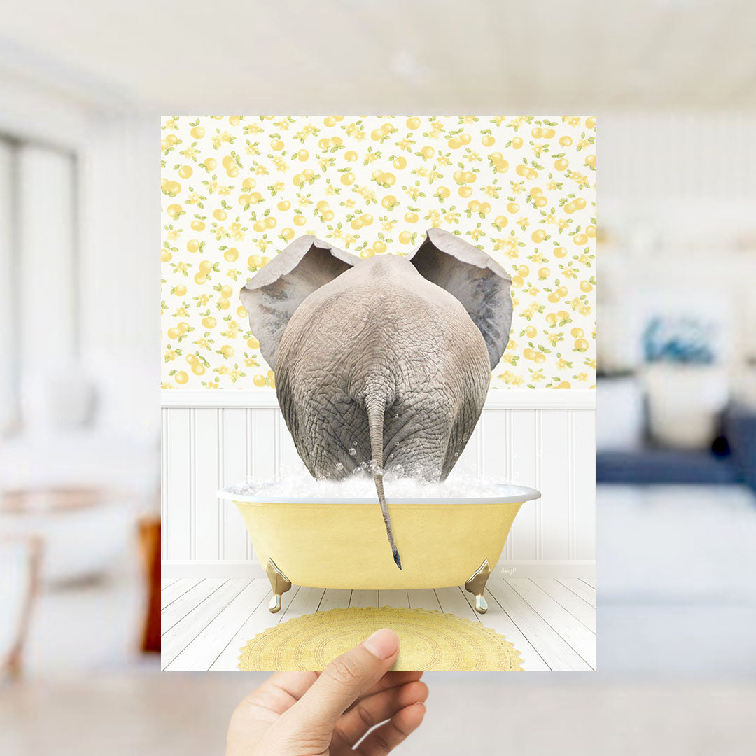 A hand holds up a framed image depicting an elephant sitting in a yellow bathtub filled with water, surrounded by a yellow and white floral pattern.
