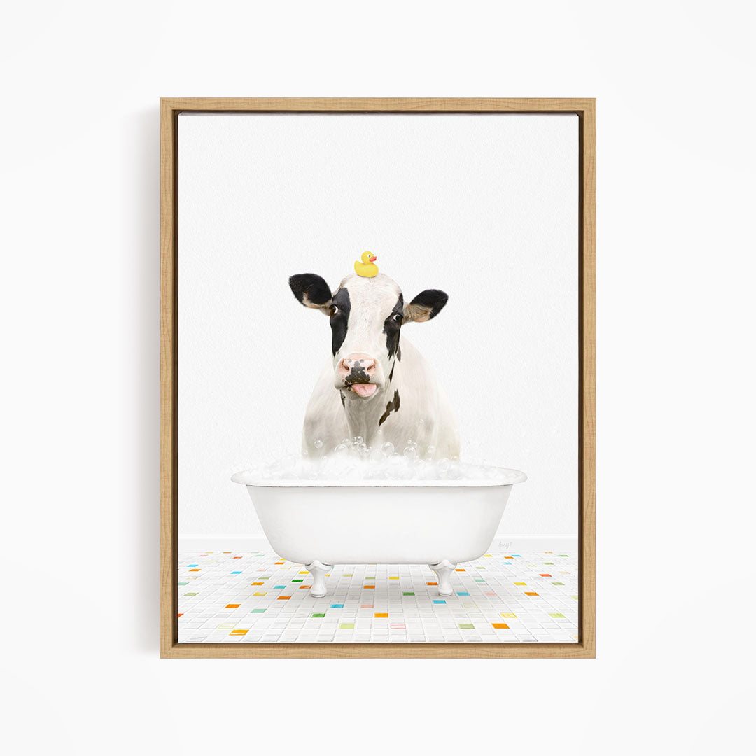 A black and white cow is sitting in a white bathtub filled with bubbles, with a yellow rubber duck perched on its head.