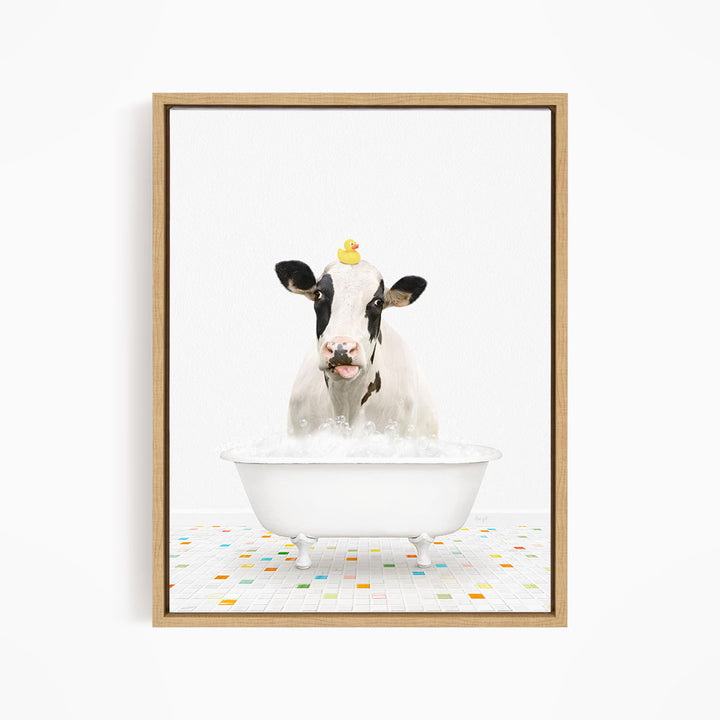 A black and white cow is sitting in a white bathtub filled with bubbles, with a yellow rubber duck perched on its head.