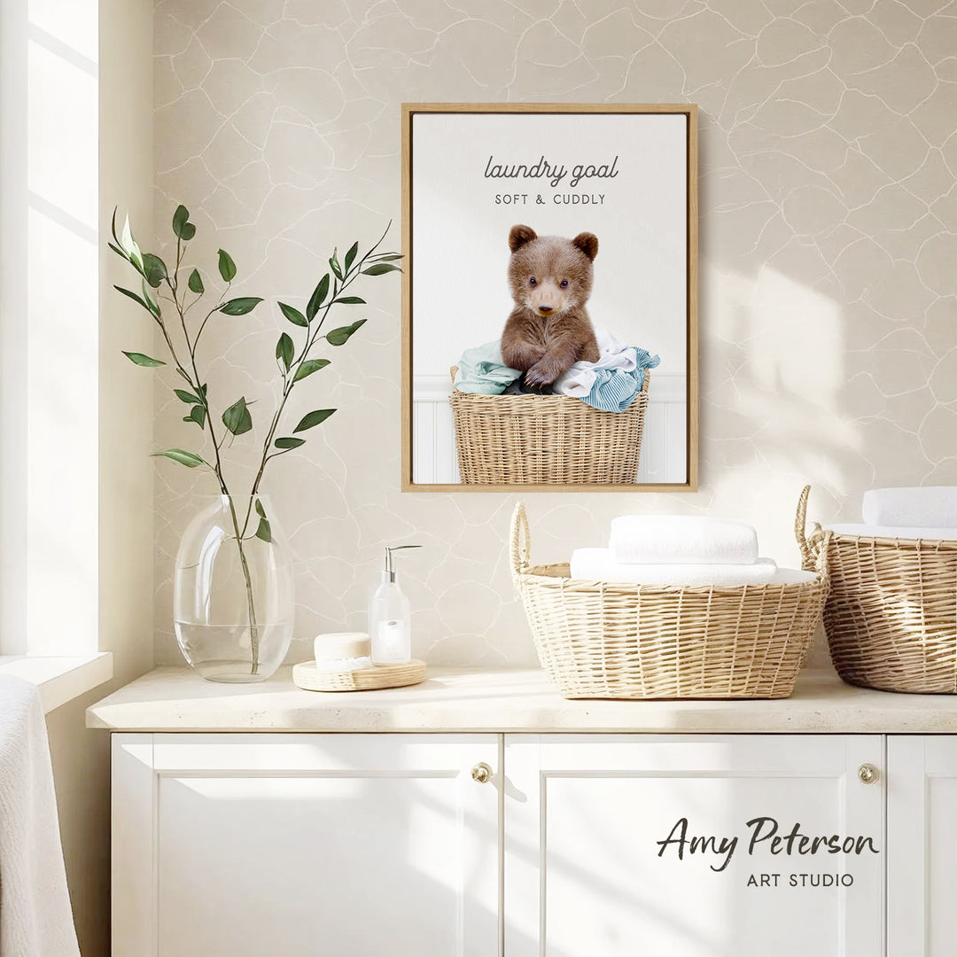 a framed photo of a baby bear sitting in a wicker basket, with the caption "laundry goal soft & cuddly". The photo is displayed on a wall above a white cabinet, which has a vase of leaves and a bottle of soap on it.
