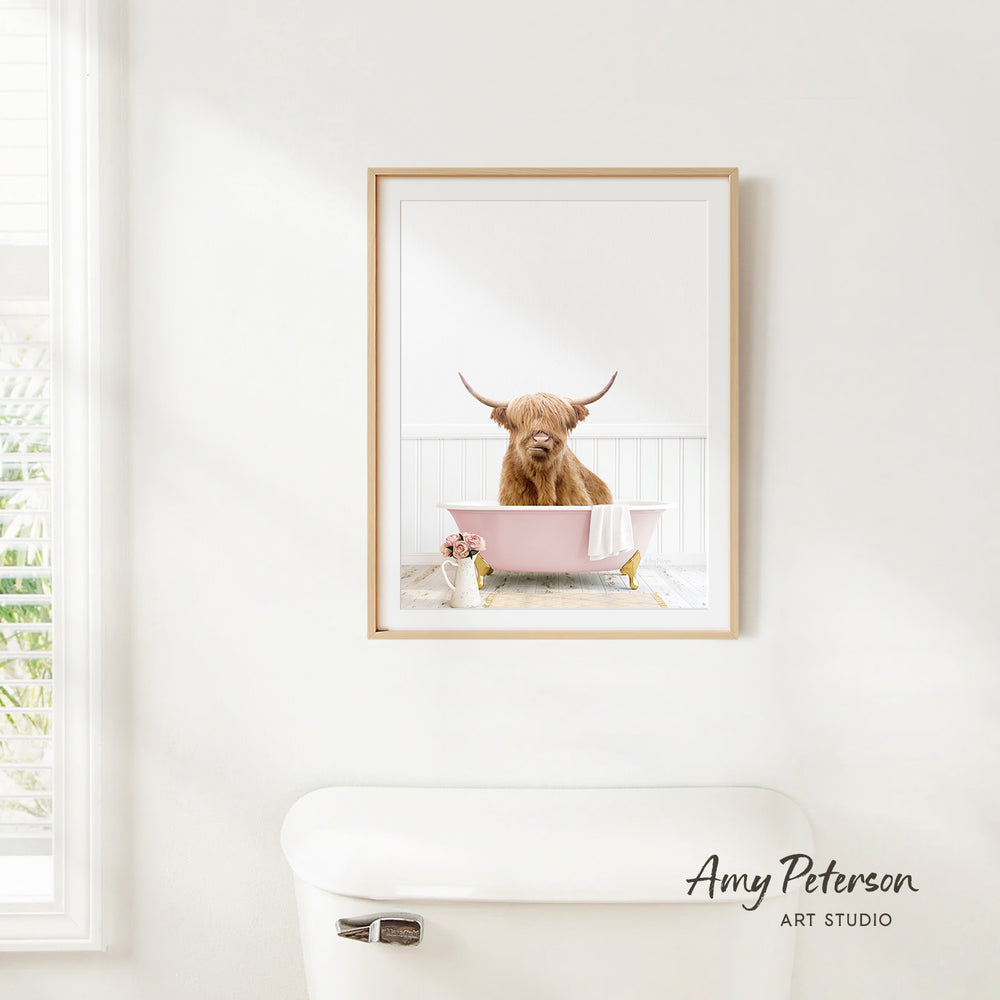 A framed image of a Highland cow sitting in a pink bathtub, with a white toilet and a window in the background.