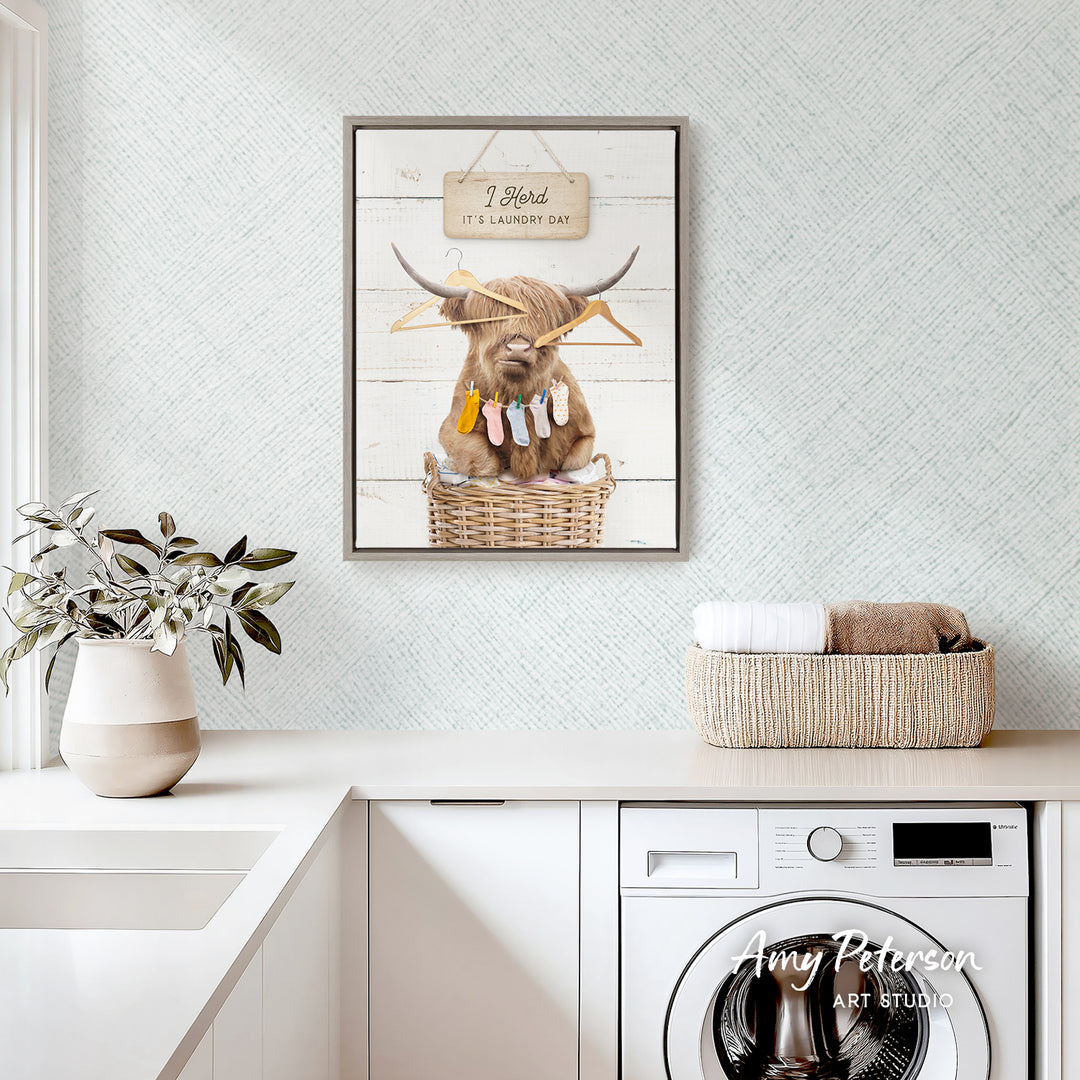 a framed picture of a Highland cow hanging on a wall above a laundry basket, with a potted plant and a basket of towels on a counter below.
