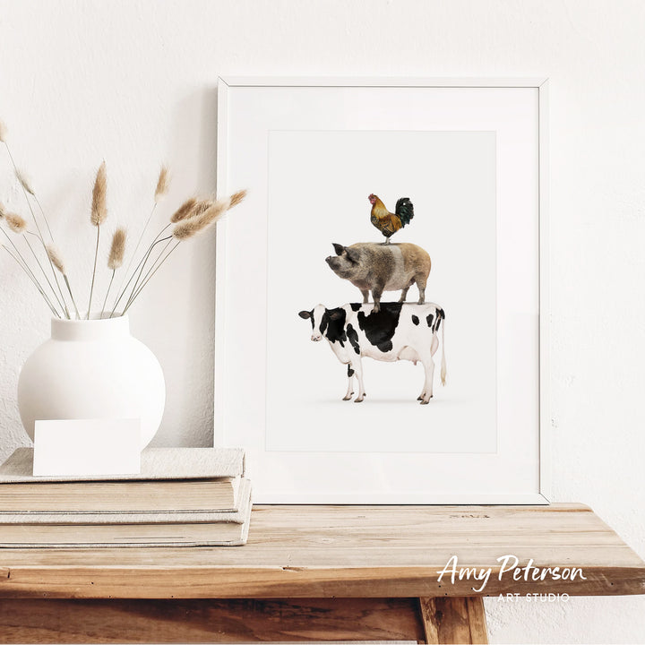 A framed illustration of a pig, cow, and rooster on a white background, placed on a wooden shelf next to a white vase with dried flowers.