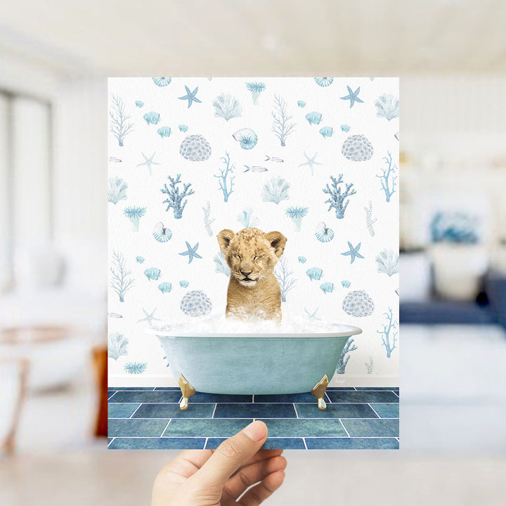 A hand is holding up a print featuring a young lion cub sitting in a bathtub filled with bubbles, surrounded by a blue and white ocean-themed wallpaper.