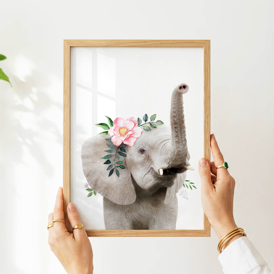 A person's hands are holding a wooden frame with a framed image of a gray elephant adorned with pink flowers and green leaves.