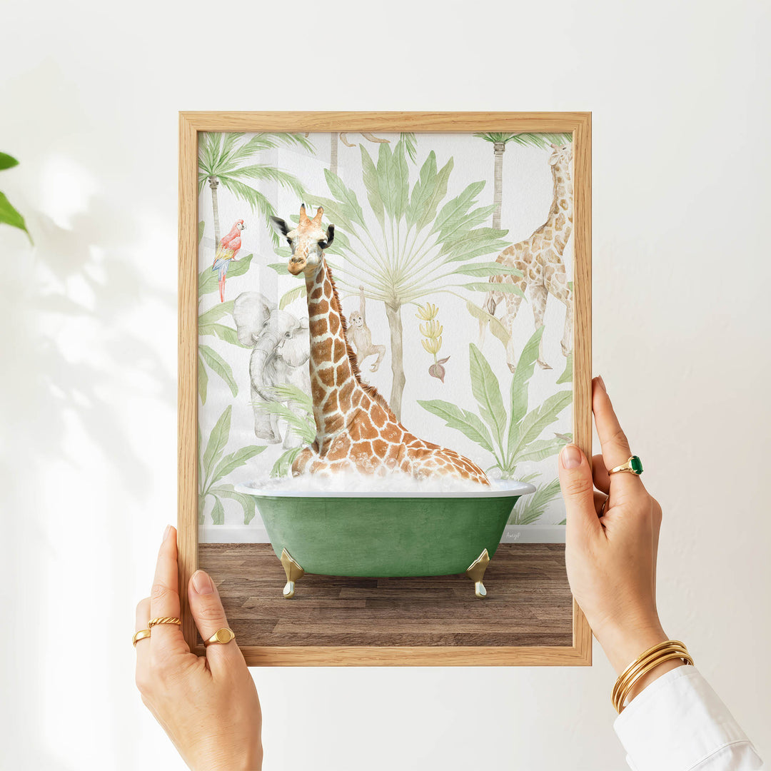 A person's hands are holding a framed picture of a giraffe sitting in a green bathtub surrounded by tropical foliage.