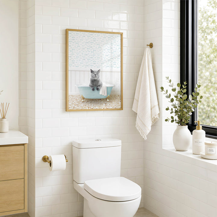 A bathroom with a white toilet, a framed picture of a cat sitting in a bathtub, and a window with greenery on the sill.