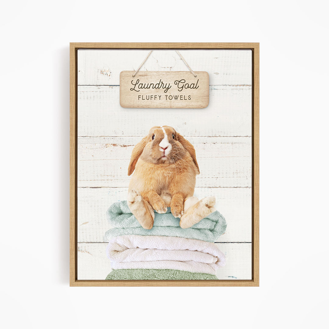 A wooden sign with the text "Laundry Goal Fluffy Towels" hangs above a stack of folded towels, with a rabbit sitting on top of the towels.