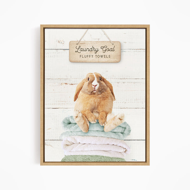 A wooden sign with the text "Laundry Goal Fluffy Towels" hangs above a stack of folded towels, with a rabbit sitting on top of the towels.