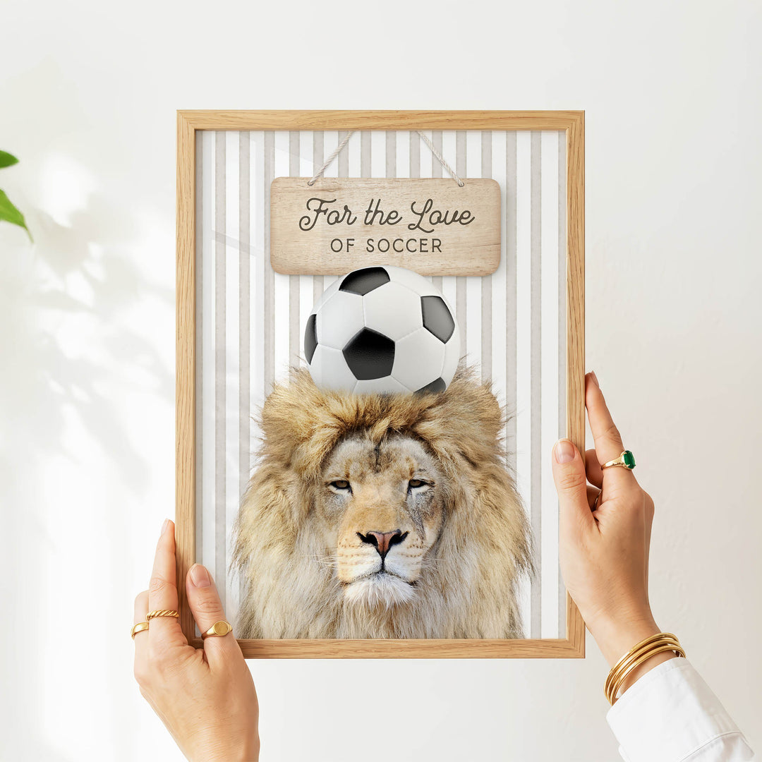 A person's hands are holding a framed picture of a lion with a soccer ball on its head, displaying a sign that reads "For the Love of Soccer".