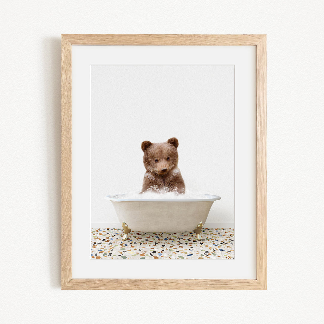 A cute brown bear cub sitting in a white bathtub filled with bubbles, surrounded by a tiled floor.