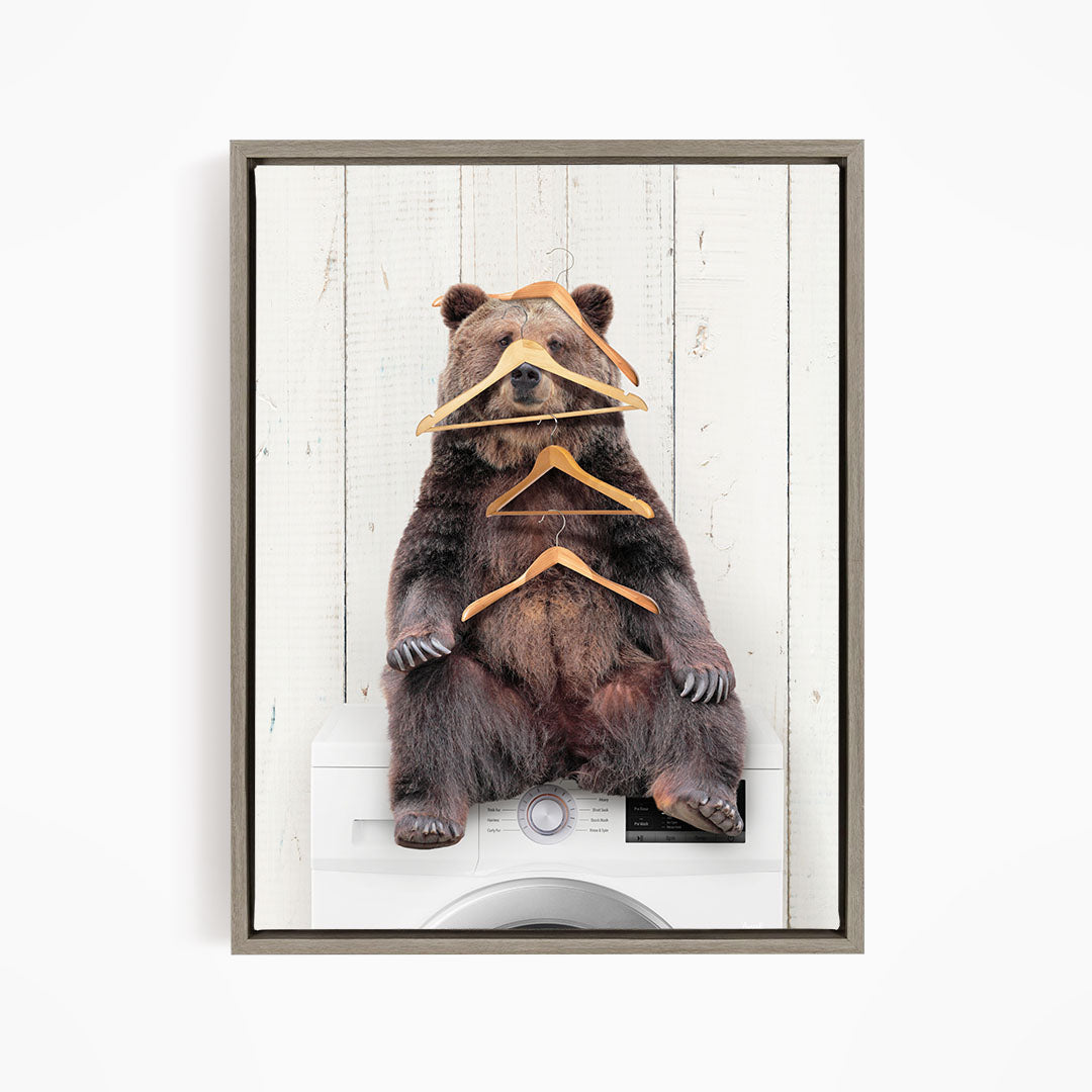 A brown bear sitting on top of a washing machine, surrounded by hanging clothes.