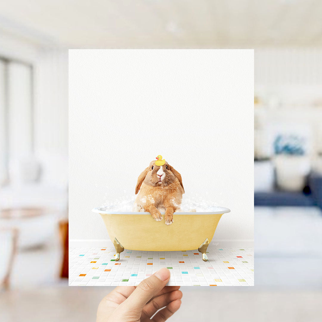 A hand holds up a photograph of a brown rabbit sitting in a yellow bathtub filled with bubbles, with a blurred background suggesting a bathroom setting.