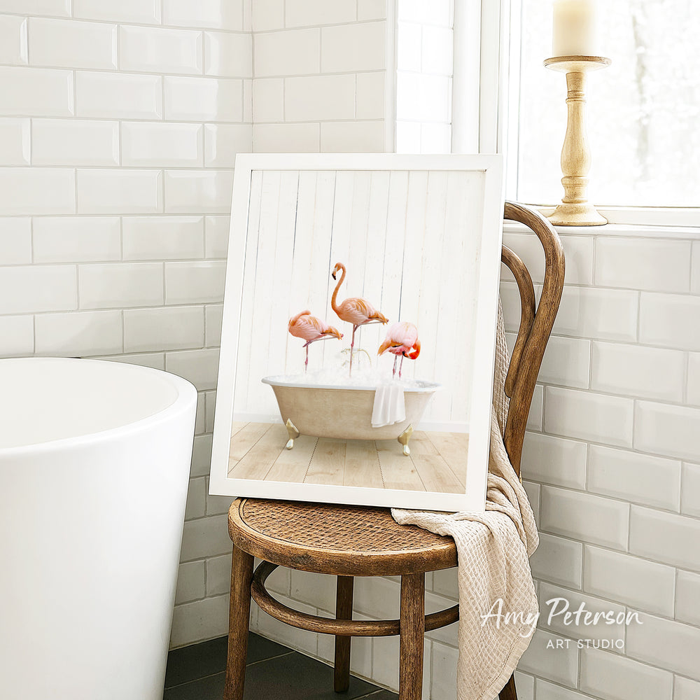 A framed image of three flamingos sitting in a bathtub is displayed on a wooden chair in a bathroom setting.