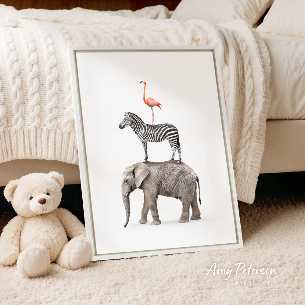 A framed print of a zebra, flamingo, and elephant is displayed on a white background, accompanied by a teddy bear and a blanket.