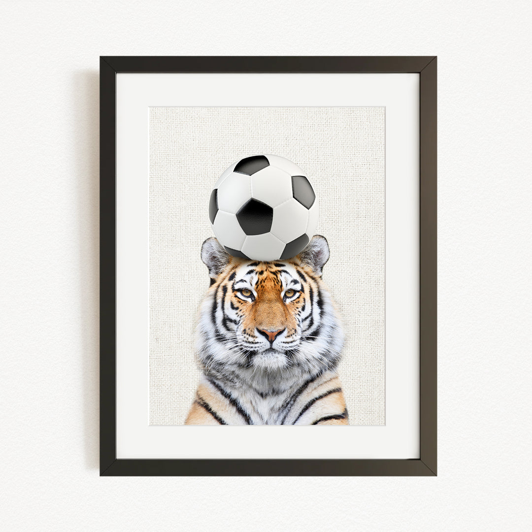 A tiger is sitting in front of a soccer ball, with the ball resting on its head.