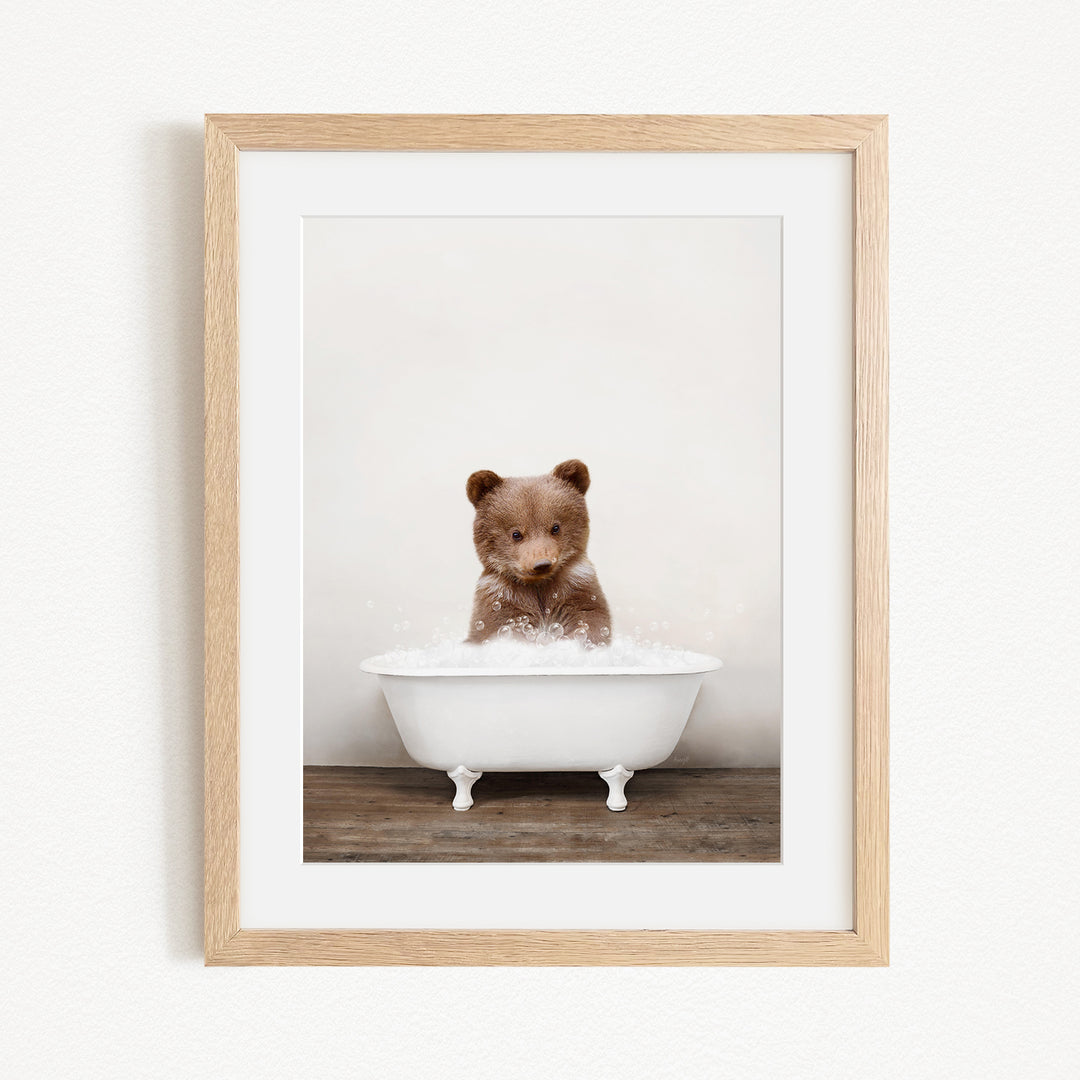 A cute brown bear sitting in a white bathtub filled with bubbles, looking directly at the camera.